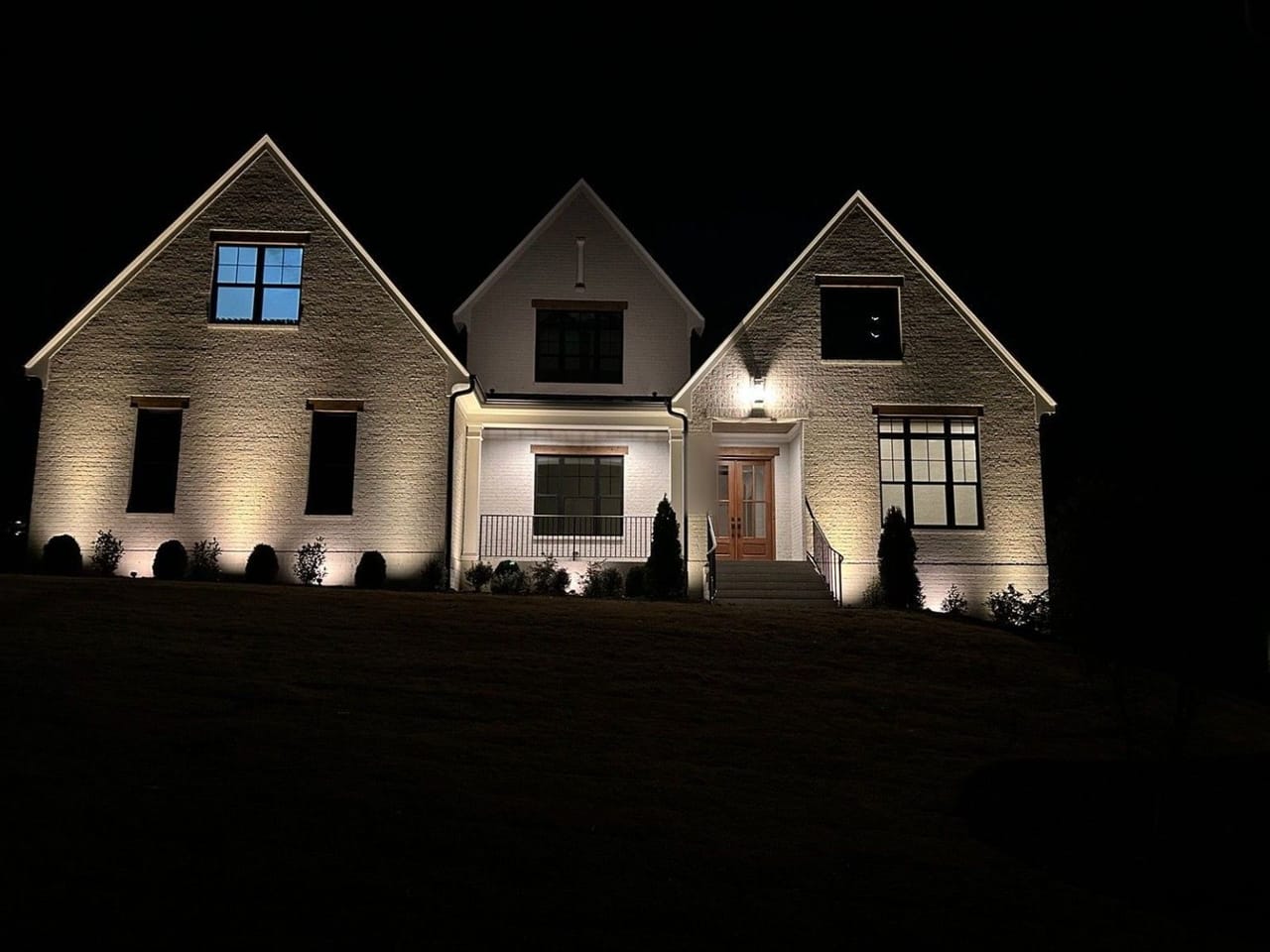 Painted brick home with three gables lit by up lights between foundation shrubs at night in Wake County