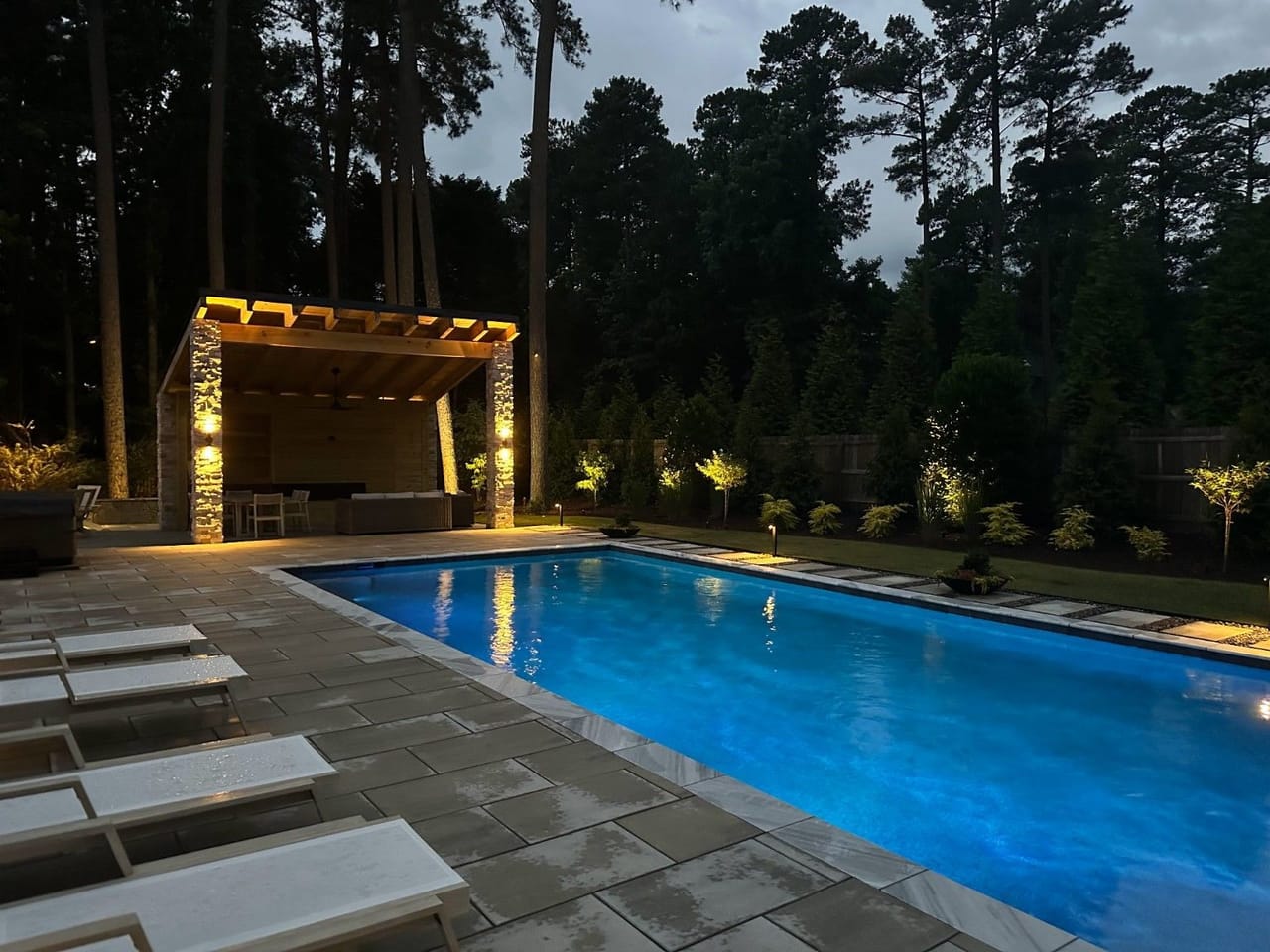 Blue-lit pool beside a stone-column pavilion with uplit trees and black path lights at dusk in Wake County
