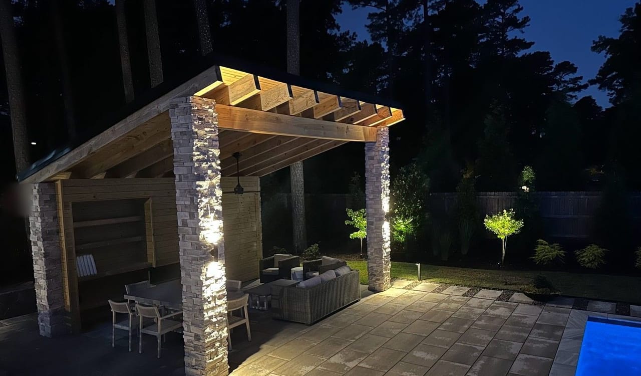 Stone-column pool pavilion with lit beams and black column sconces beside uplit trees at night in Wake County